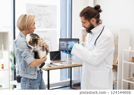 Veterinarian explaining X-ray image to young woman holding her pet cat indoors office. Image suitable for health care, veterinarian services, and animal welfare. 124144608