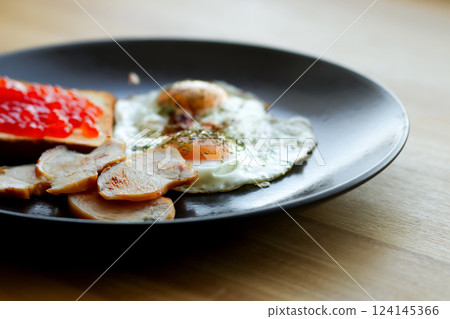 a delicious American-style breakfast plate featuring a fried egg, crispy bacon, and a touch of red caviar. American breakfast on the black plate close up a delicious American-style breakfast plate featuring a fried egg, crispy bacon, and a touch of red caviar. American breakfast on the black plate close up 124145366