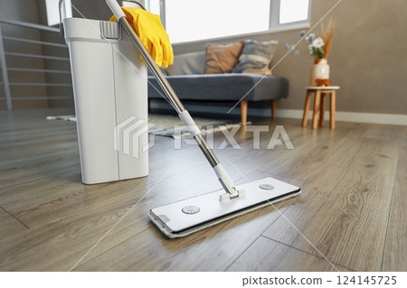 Cleaning supplies: mop, gloves, bucket with wringer sitting on wooden laminate floor in modern living room, wet equipment tools, housekeeping concept. Emphasizing home sanitation, cleanliness, hygiene 124145725