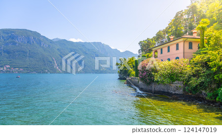 A charming villa overlooks the tranquil waters of Lake Como, surrounded by lush greenery and majestic mountains under a clear blue sky, inviting visitors to enjoy the picturesque scenery. A charming villa overlooks the tranquil waters of Lake Como, surrounded by lush greenery and majestic mountains under a clear blue sky, inviting visitors to enjoy the picturesque scenery. 124147019