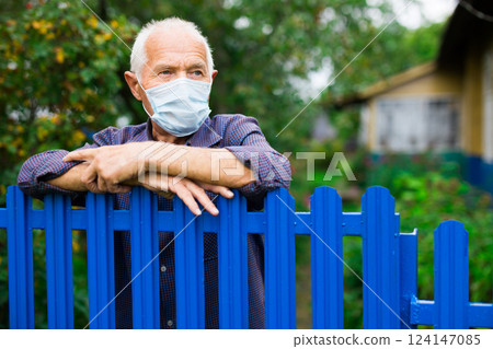Portrait of elderly man in protective mask at fence of his country house in the village. Isolation concept during pandemic Portrait of elderly man in protective mask at fence of his country house in the village. Isolation concept during pandemic 124147085