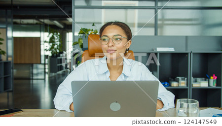 A young professional woman wearing glasses working on a laptop, sitting in a modern office with shelves, plants, and natural light in the background A young professional woman wearing glasses working on a laptop, sitting in a modern office with shelves, plants, and natural light in the background 124147549