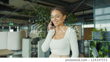 Smiling professional woman on a phone call in a bright, modern office surrounded by greenery, sleek furniture, and natural light, exuding confidence and joy. Smiling professional woman on a phone call in a bright, modern office surrounded by greenery, sleek furniture, and natural light, exuding confidence and joy. 124147553