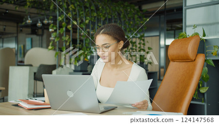 A focused professional woman analyzing documents and working on a laptop in a modern, green office space with stylish furniture, productivity tools, and natural light A focused professional woman analyzing documents and working on a laptop in a modern, green office space with stylish furniture, productivity tools, and natural light 124147568