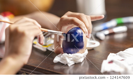 A little girl is painting on an egg with a paintbrush for easter egg decoration on the table. Concept of celebrating the easter holiday A little girl is painting on an egg with a paintbrush for easter egg decoration on the table. Concept of celebrating the easter holiday 124147609