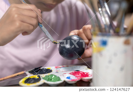 Indonesian southeast asian little girl is painting on an egg with a paintbrush for easter egg decoration on the table. Concept of celebrating the easter holiday Indonesian southeast asian little girl is painting on an egg with a paintbrush for easter egg decoration on the table. Concept of celebrating the easter holiday 124147627
