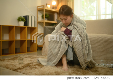 Sick woman sitting on the floor wrapped in a gray blanket. Illness and home recovery concept Sick woman sitting on the floor wrapped in a gray blanket. Illness and home recovery concept 124148689