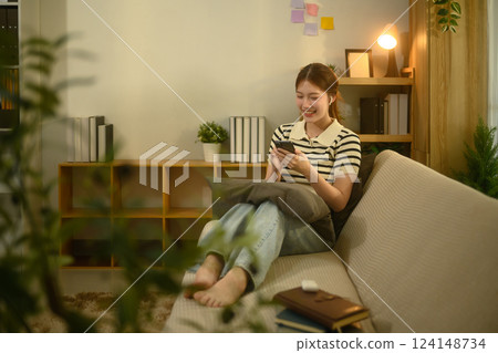Smiling young asian woman enjoying social media on smartphone sitting on couch at home 124148734