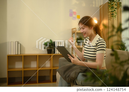 Cheerful young woman sitting on couch raising her fist in excitement while looking at her digital tablet 124148754