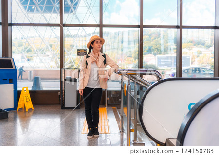 Pretty Caucasian elegant young woman in straw hat posing near escalator. Windows in background. The concept of vacation and flight trip 124150783