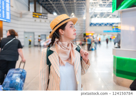 Young Caucasian elegant pretty woman in straw hat, wearing backpack, holds her passport to check in for a flight. Waiting in airport 124150784