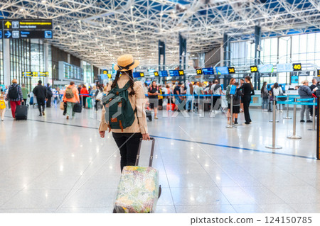 Back view of elegant woman in a straw hat and backpack, wheeling a suitcase to the check-in desk. Copy space. The concept of vacation and flight trip 124150785