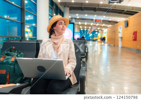 Young Caucasian elegant happy woman is sitting on a chair in waiting room of airport and working on a laptop. Copy space. Concept of remote work and travel 124150798