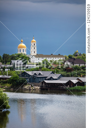 A picturesque town with a river in the foreground, lush greenery, and golden-domed churches in the background. The dramatic sky adds contrast, creating a serene yet moody atmosphere. 124150919