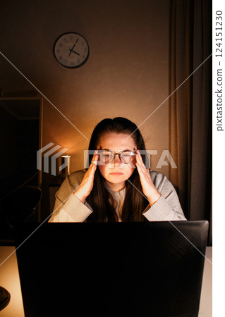 Woman working on laptop late evening at home. Concentrated female employee solves work issues sitting at computer at work desk. 124151230