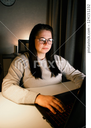 Young woman with eyeglasses working on laptop at home late at night, using modern technologies and getting online education. Remote learning concept. Online university. Focus on process. 124151232