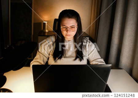 Woman working on laptop late evening at home. Concentrated female employee solves work issues sitting at computer at work desk. 124151248