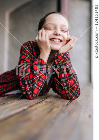 Joyful 10 years old girl with braided hair, wearing red checkered shirt, with eyes closed, smiling and holding face with hands, conveying sense of joy and happiness, leaning on rustic wooden table. 124151483