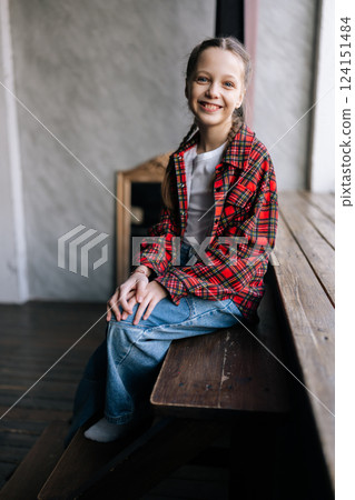 Vertical portrait of pretty ten-year-old girl with braided hair wearing red plaid shirt and jeans, sitting on wooden bench by window at day, enjoying relaxed moment indoors, smiling looking at camera. Vertical portrait of pretty ten-year-old girl with braided hair wearing red plaid shirt and jeans, sitting on wooden bench by window at day, enjoying relaxed moment indoors, smiling looking at camera. 124151484