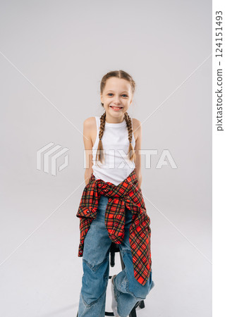 Studio vertical portrait of joyful 10 years old girl with braided hair posing smiling looking at camera. Pretty preteen child wearing white tank top, blue jeans, red checkered shirt knotted at waist. Studio vertical portrait of joyful 10 years old girl with braided hair posing smiling looking at camera. Pretty preteen child wearing white tank top, blue jeans, red checkered shirt knotted at waist. 124151493