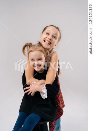 Vertical portrait of two young sisters embracing and laughing, creating heartwarming portrait of sibling love and happiness in professional studio setting with white background, looking at camera. 124151526