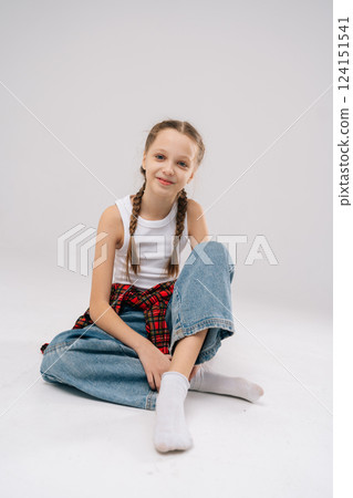 Vertical portrait of pretty young girl with braided hair wearing white tank top and red plaid shirt, sitting cross-legged with cheerful expression against neutral backdrop, smiling looking at camera, Vertical portrait of pretty young girl with braided hair wearing white tank top and red plaid shirt, sitting cross-legged with cheerful expression against neutral backdrop, smiling looking at camera, 124151541