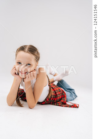 Vertical portrait of pretty 10 years old girl with braided hair in white tank top, red plaid shirt, lying on white backdrop, resting chin on hands, looking at camera. Concept of carefree childhood. Vertical portrait of pretty 10 years old girl with braided hair in white tank top, red plaid shirt, lying on white backdrop, resting chin on hands, looking at camera. Concept of carefree childhood. 124151546