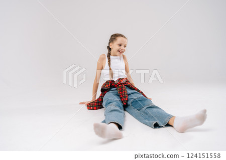 Studio portrait of cheerful preteen girl sitting on white floor with checkered shirt tied around waist, showcasing casual and playful style. Concept of carefree childhood. 124151558