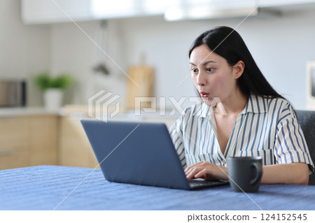 Surprised asian woman using laptop in the kitchen Surprised asian woman using laptop in the kitchen 124152545