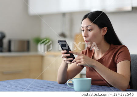 Surprised asian woman using smart phone in a kitchen Surprised asian woman using smart phone in a kitchen 124152546