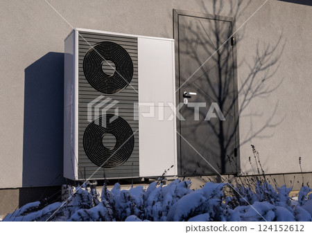 Modern heat pump unit installed outside a residential building in winter, surrounded by snow-covered plants. Energy-efficient heating solution in cold weather. 124152612