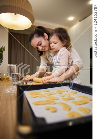 Happy mother and cute little child daughter rolling dough for homemade cookies and tray with gingerbread cookies Happy mother and cute little child daughter rolling dough for homemade cookies and tray with gingerbread cookies 124152787