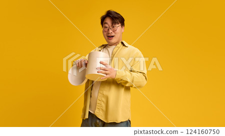 Young Asian man, dressed casually, get present and looking surprised while unpack it against sunny yellow background. 124160750