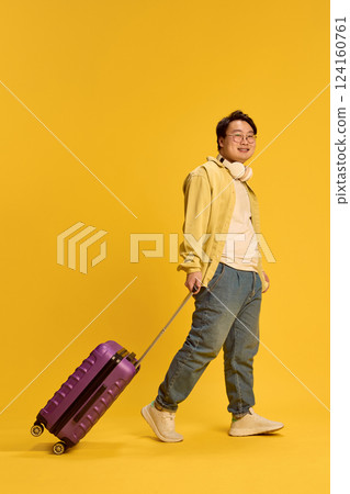 Cheerful smiling young Asian man walking, dressed casual attire and headphones, with luggage, ready to trip against sunny yellow background. Cheerful smiling young Asian man walking, dressed casual attire and headphones, with luggage, ready to trip against sunny yellow background. 124160761