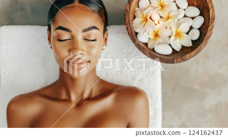 Spa day relaxation with a serene young woman resting with closed eyes on a soft towel beside a wooden bowl filled with frangipani flowers and white stones. 124162437