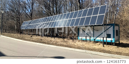 A solar panel array stands next to a small utility building in a wooded area. Sunlight reflects off the panels, emphasizing the commitment to renewable energy 124163773