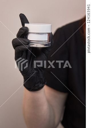 Hand in black glove holding white cosmetic jar, closeup of beauty product in professional care setting, skincare focus concept Hand in black glove holding white cosmetic jar, closeup of beauty product in professional care setting, skincare focus concept 124163941