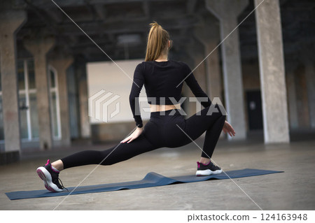 Woman in athletic attire stretching on yoga mat in industrial space, fitness and flexibility demonstration concept 124163948
