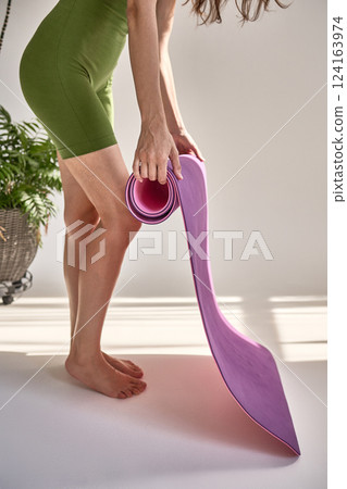 Woman in green shorts rolling pink yoga mat, preparing for indoor workout, exercise, fitness activity, healthy lifestyle concept 124163974