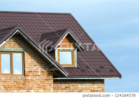 Close-up detail of new brick house top with brown shingle roof and plastic attic windows on bright blue sky background. Real estate property and professionally done work concept. Close-up detail of new brick house top with brown shingle roof and plastic attic windows on bright blue sky background. Real estate property and professionally done work concept. 124164576