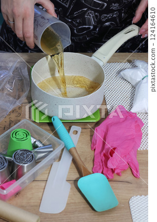 A woman is pouring apple juice into a saucepan. She is preparing syrup for homemade marshmallows. Nearby on the table are zefir-making tools. Pastry tips for creating marshmallow flowers. A woman is pouring apple juice into a saucepan. She is preparing syrup for homemade marshmallows. Nearby on the table are zefir-making tools. Pastry tips for creating marshmallow flowers. 124164610