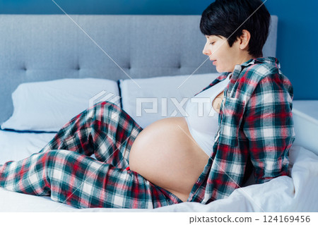 Pregnant woman sitting on bed, touching her belly, showcasing joy and anticipation. Expecting mother relaxing in pajama during last trimester of pregnancy. Motherhood, preparation and expectation 124169456