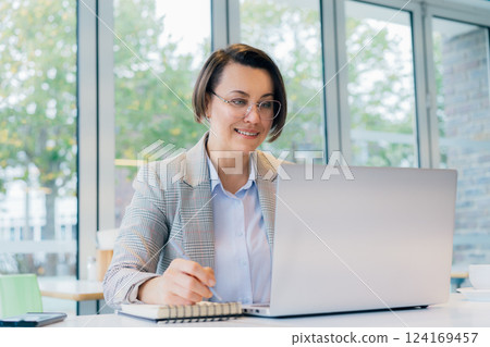 Portrait of 50's confident mature businesswoman, middle-aged experienced senior female professional making notes, working on laptop in office or cafe. Female CEO, female entrepreneur manages business 124169457