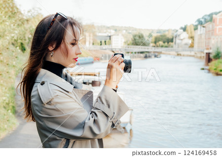 Smiling young woman holding retro style camera. Female traveller watching photos on camera during city walk. Travel blogger, photographer, influencer capture the moment. Hobby, travel concept. 124169458