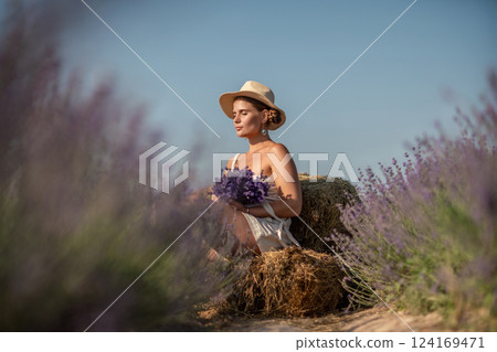 woman sitting in a field of lavender and wearing a straw hat. She is smiling and holding a bouquet of flowers. Scene is peaceful and serene, as the woman is surrounded by the beauty of nature 124169471