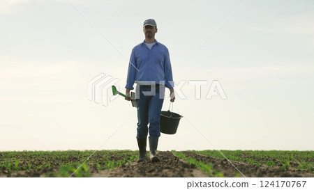 Farmer with shovel bucket walks through agricultural field. Male worker employee going to manually prepare soil for sowing planting cultivated plants. Loosening weed removal soil structure improvement 124170767