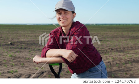 Woman farmer portrait with shovel. Young happy smiling village woman in field leaning on handle of shovel looking at camera with satisfied delighted vid, enjoying manual labor work in agrarian field. 124170768