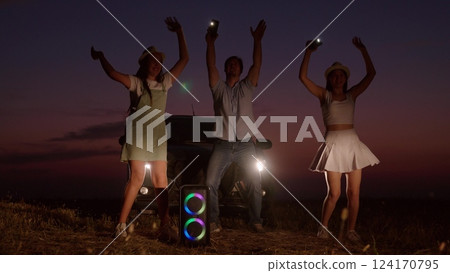 Cheerful company dances by car in evening. People friendly best close friends family on road trip stopped at night jumping partying to music next to glowing rainbow music column with flashlights. Cheerful company dances by car in evening. People friendly best close friends family on road trip stopped at night jumping partying to music next to glowing rainbow music column with flashlights. 124170795