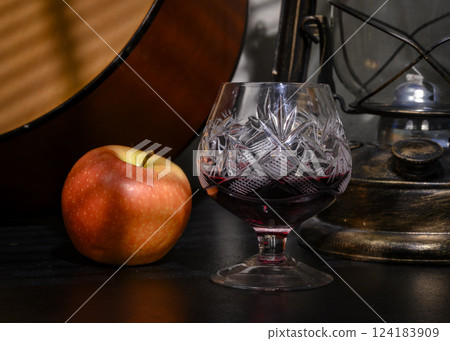 Old crystal glass with red wine on the background of an apple, a bronze kerosene lamp and a guitar 124183909