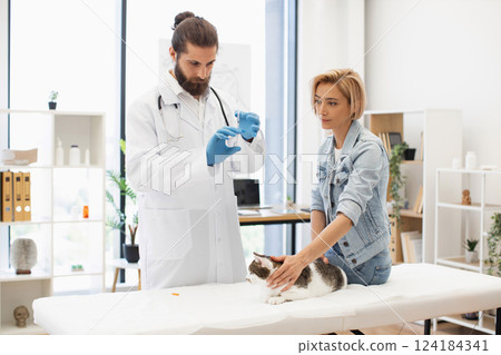 Male veterinarian administering injection to cat as young female owner holds the pet. 124184341